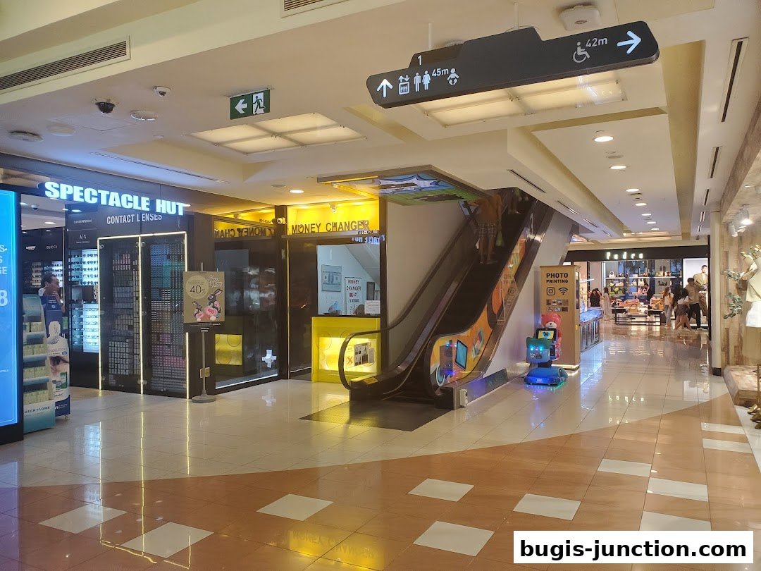 A shopping mall interior with a money changer, spectacle shop, and escalator.