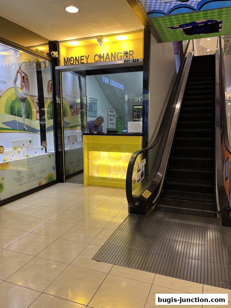 A money changer shop with a bright yellow counter and signage, located next to an escalator.