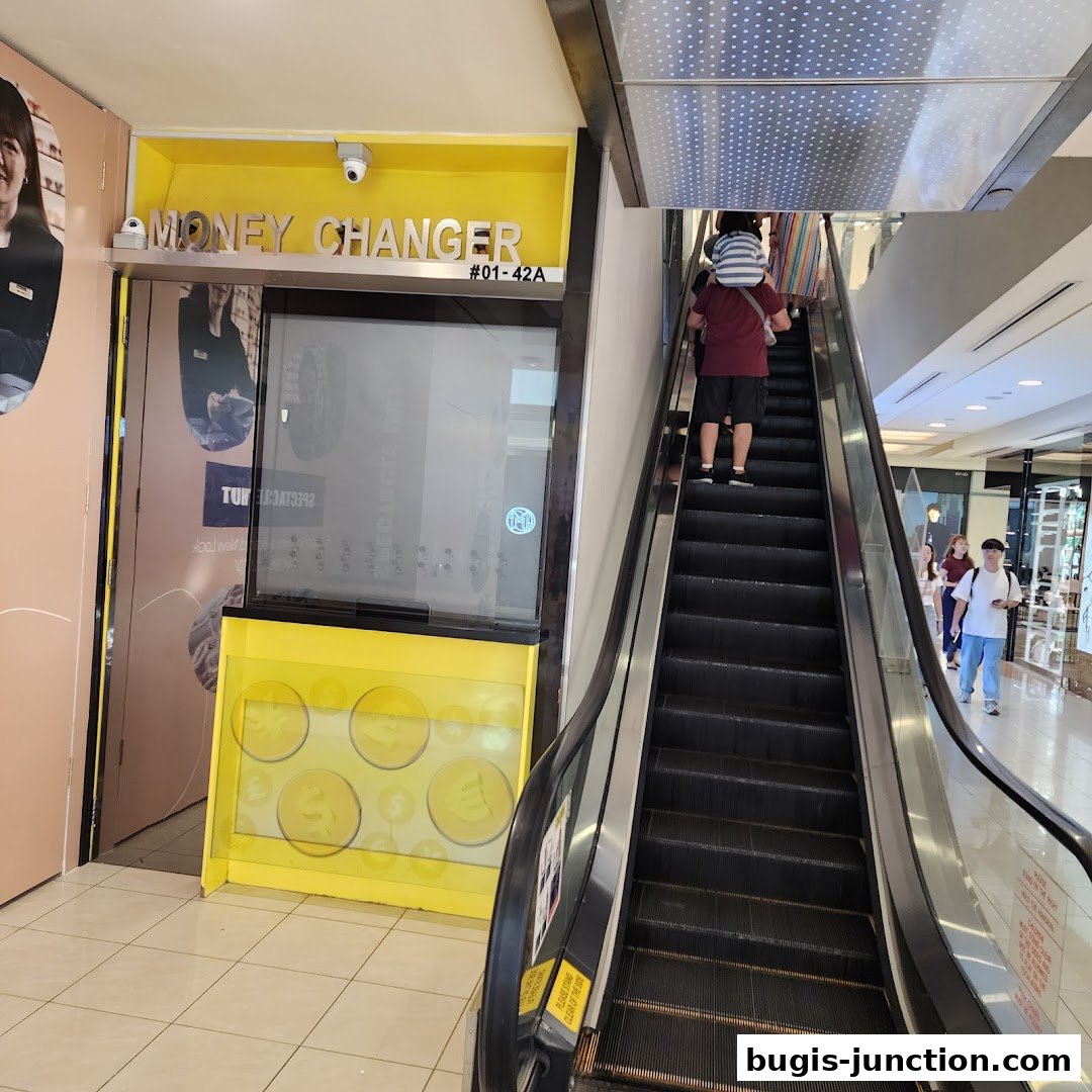 The exterior of a brightly lit money changer shop with a prominent yellow sign and an escalator nearby.
