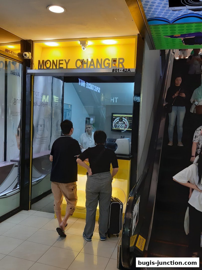 A money changer shop with customers interacting with staff, located near an escalator.
