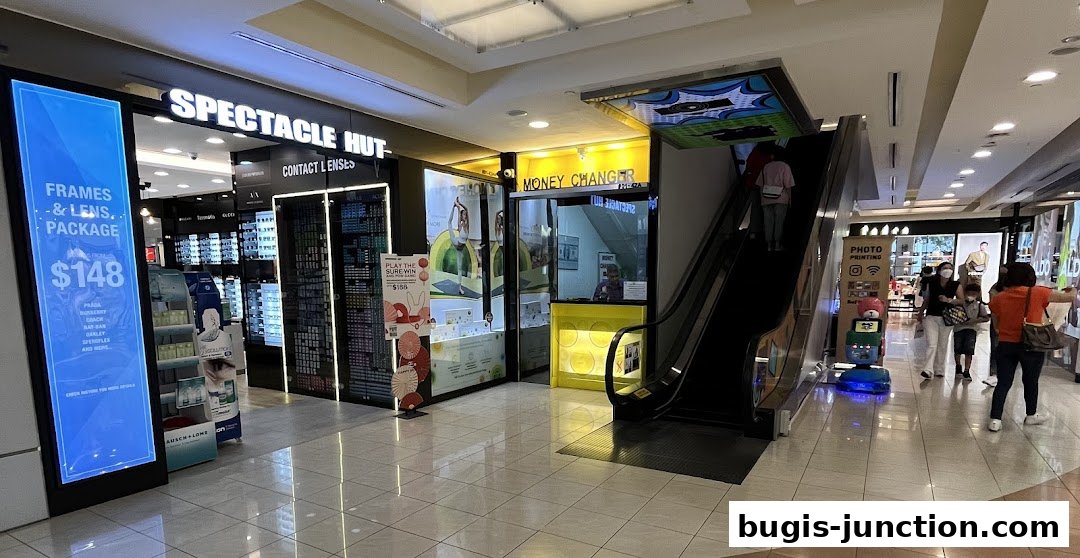 A shopping mall corridor with shops including Spectacle Hut and a Money Changer, plus an escalator.