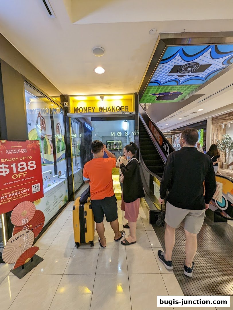 People stand outside a money changer shop with a yellow sign and a display window.