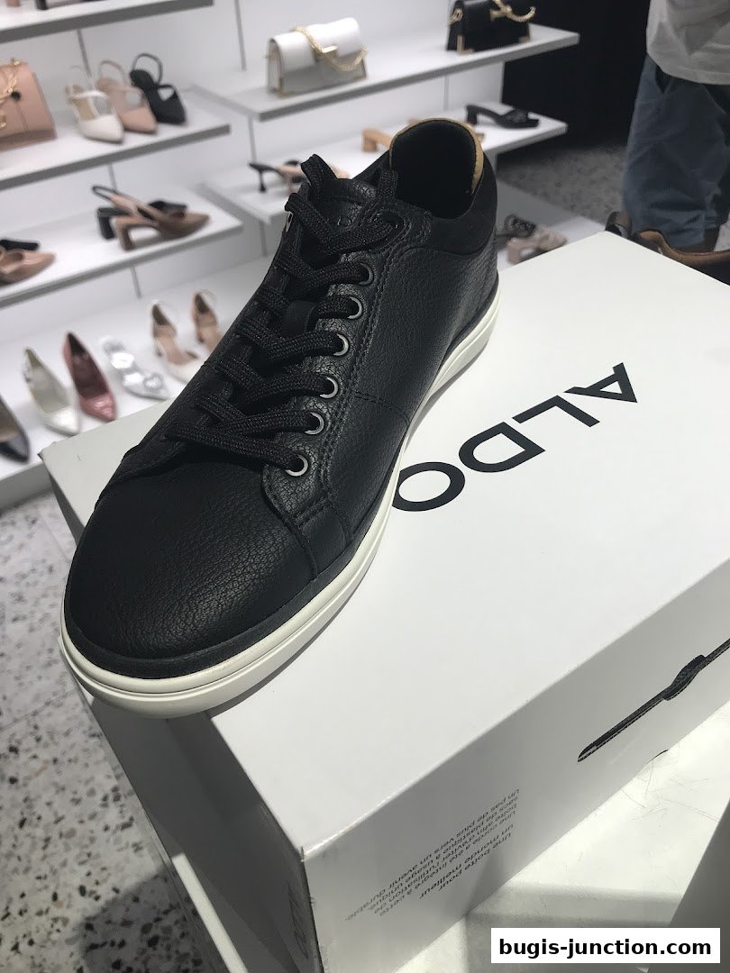 A black ALDO sneaker is displayed on a white ALDO shoebox, with shelves of shoes and bags in the background.