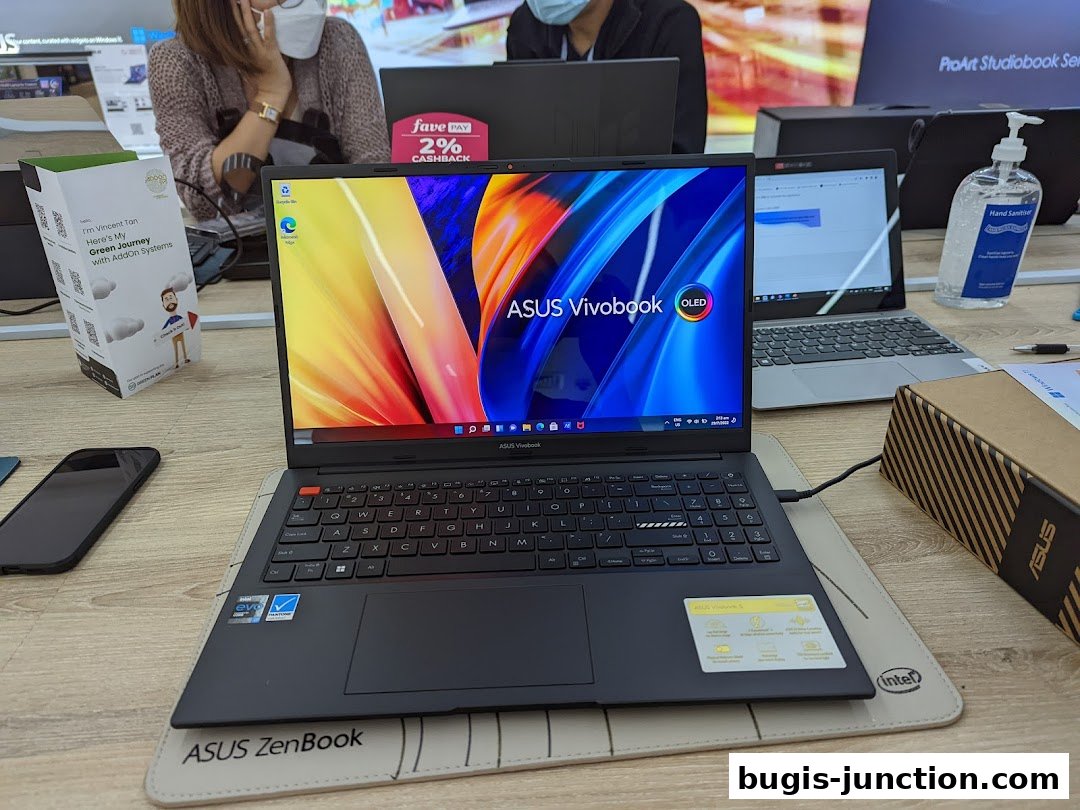 An ASUS Vivobook laptop is displayed on a wooden desk with other laptops and hand sanitizer.