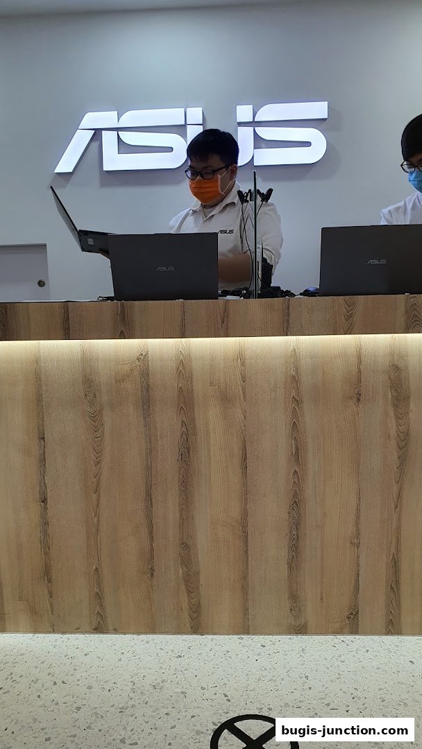 Two staff members wearing masks stand behind a counter with ASUS laptops and branding.