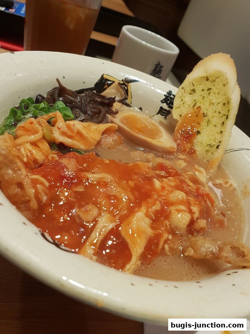 A close-up of a ramen bowl with toppings, a slice of garlic bread, and a drink.