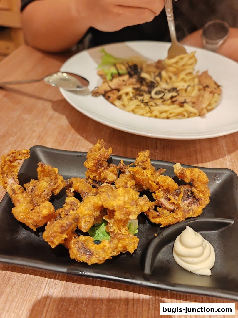 A plate of crispy fried calamari with a side of mayonnaise and a pasta dish in the background.
