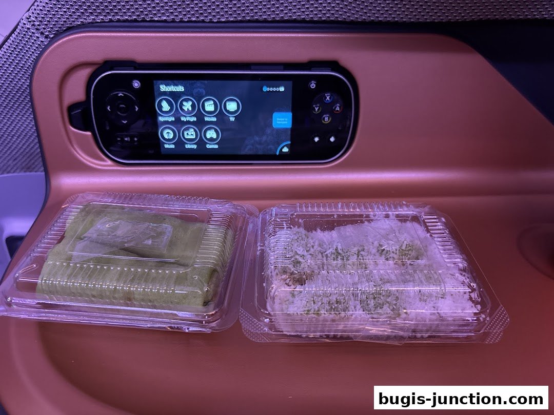 Two plastic containers of traditional Indonesian snacks on a tray with an entertainment system.