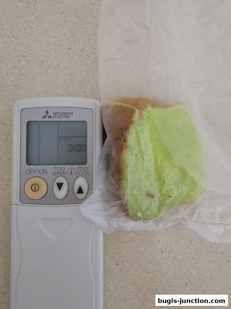 A slice of green pandan cake is wrapped in plastic next to an air conditioner remote.