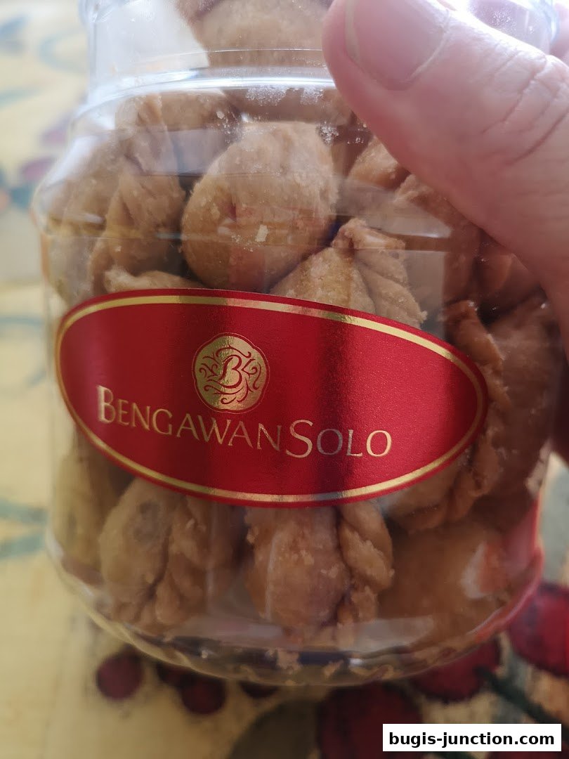A hand holds a clear jar filled with small, golden-brown pastries, branded with a red and gold label.