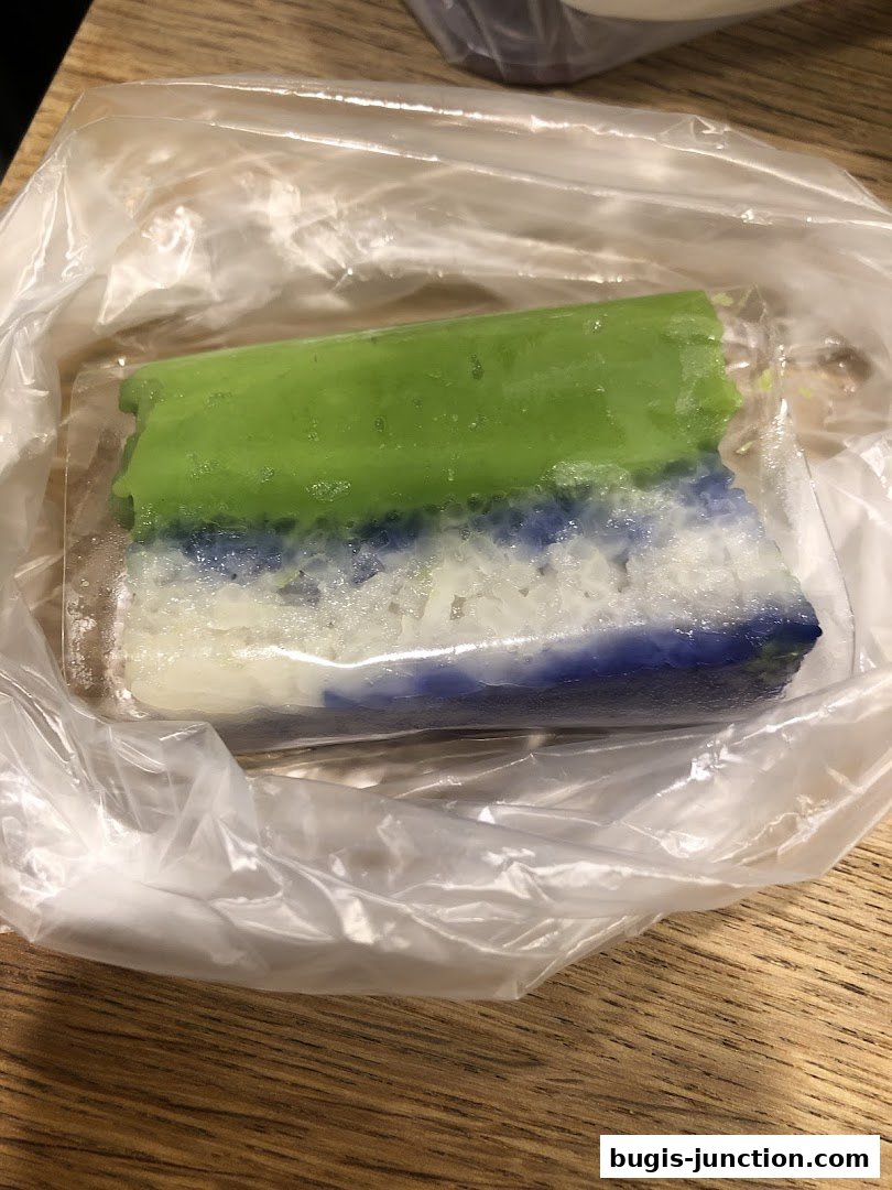 A colorful layered dessert, likely kueh, wrapped in plastic on a wooden surface.