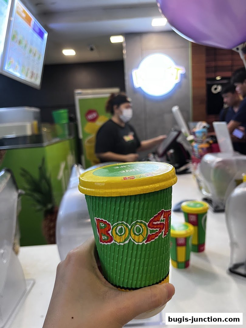 A hand holds a Boost Juice cup inside the store with staff and other cups visible.