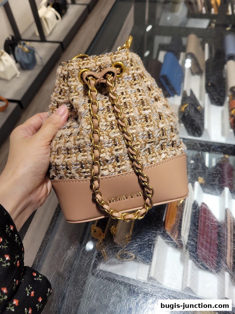 A hand holds a textured bucket bag with a chain strap in front of a display of handbags.