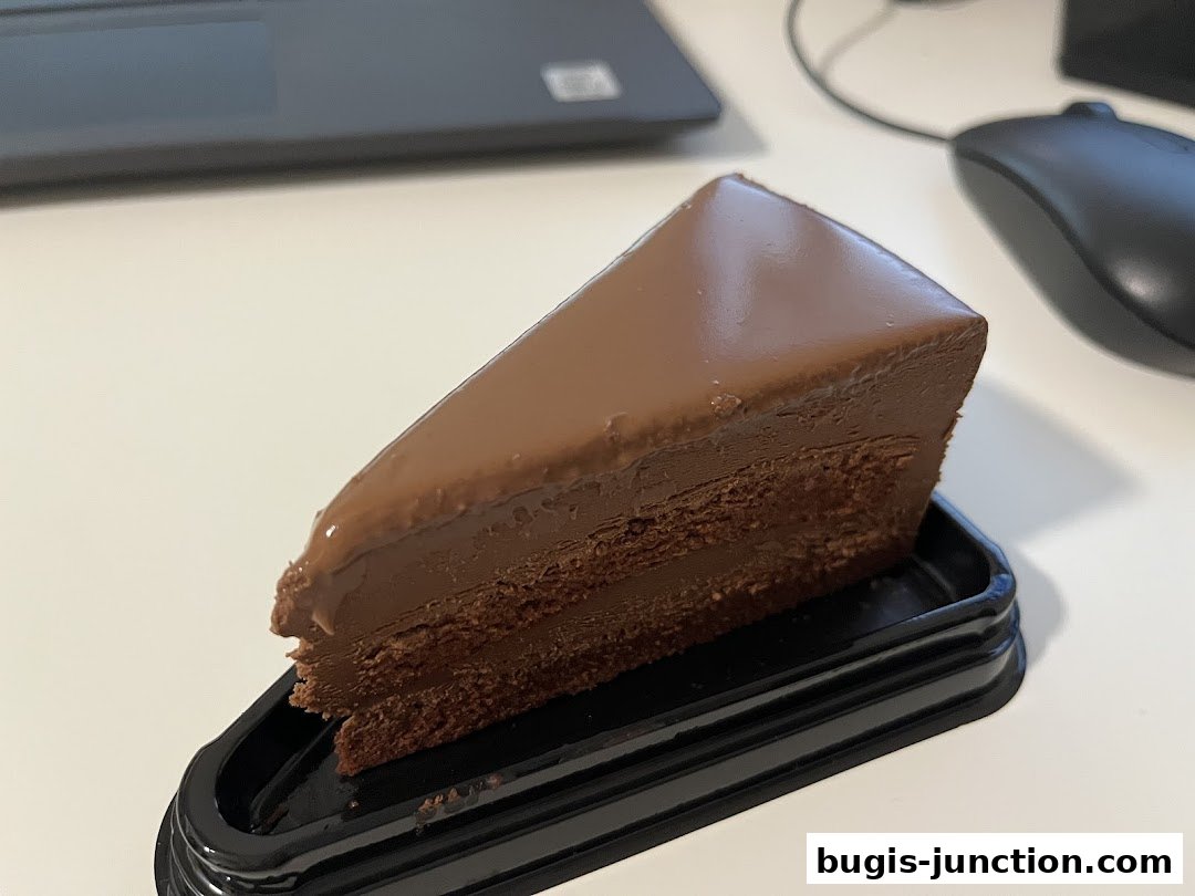 A slice of rich chocolate cake with glossy frosting on a black tray.