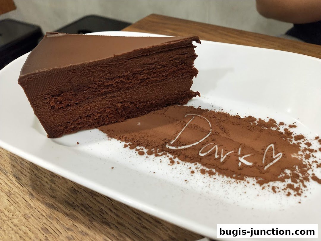 A slice of rich chocolate cake is presented on a white plate with cocoa powder.