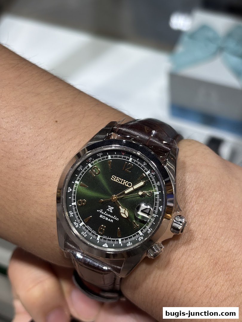 A close-up shot of a Seiko automatic watch with a green dial and brown leather strap on a wrist.