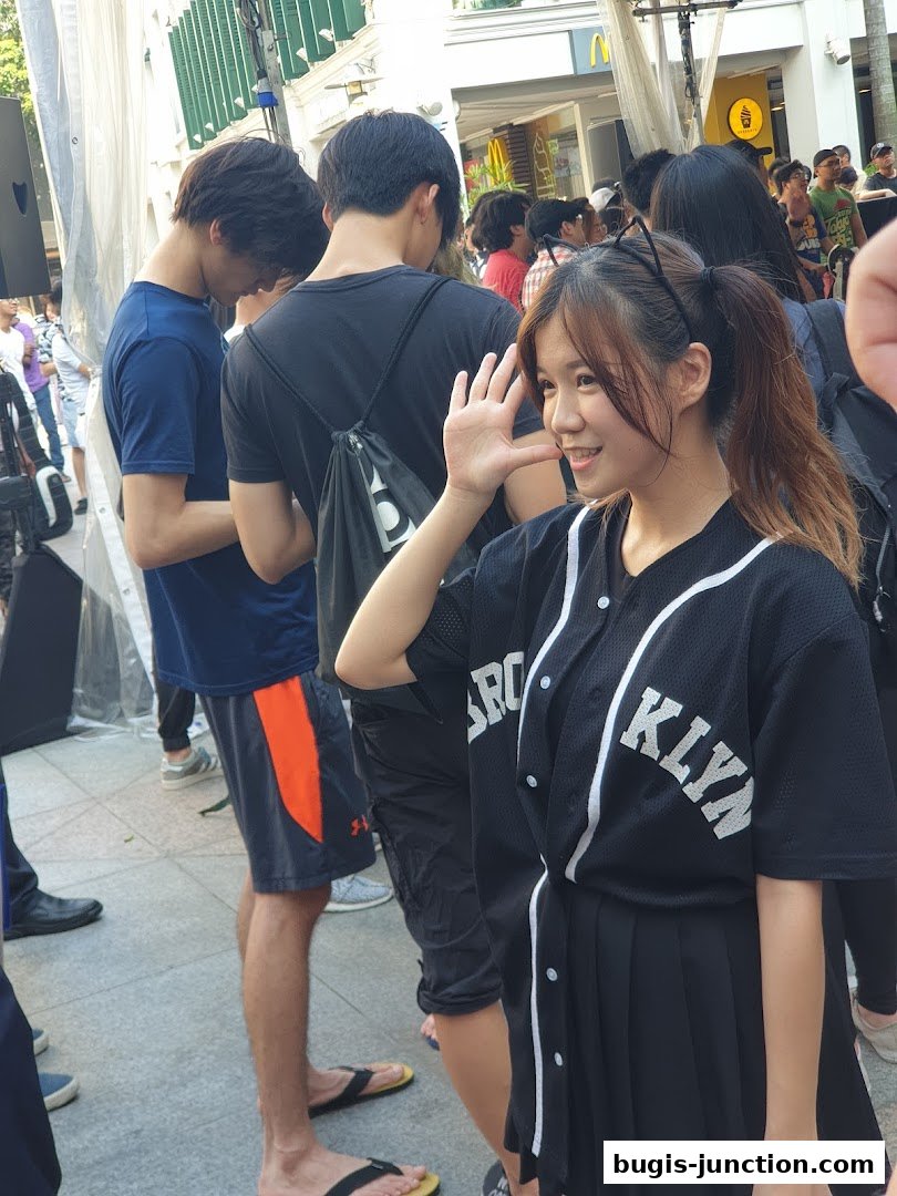 A young woman in a baseball jersey and skirt waves at the camera, surrounded by a crowd.