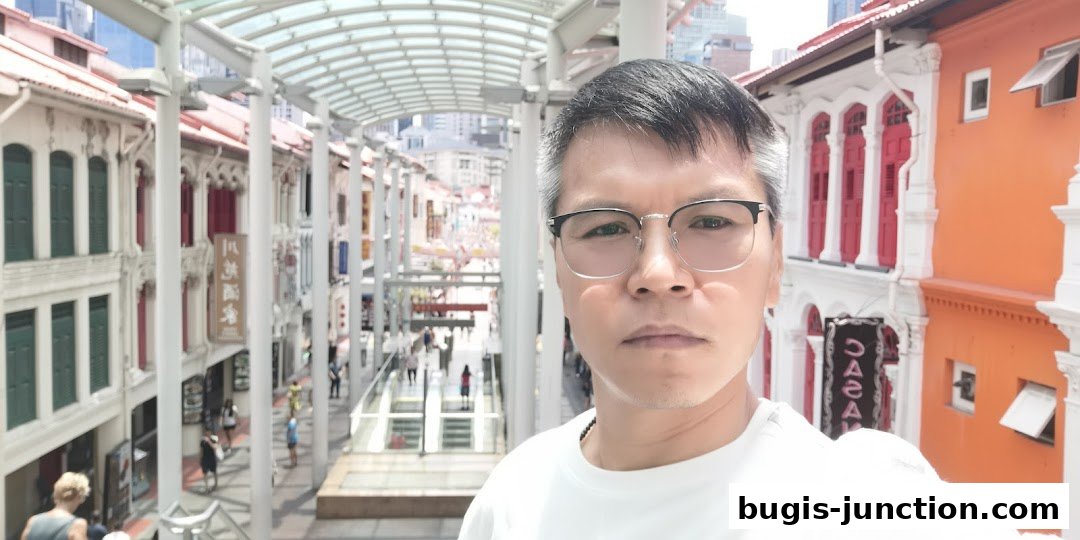 A man poses for a selfie with a street of colorful shops in the background.