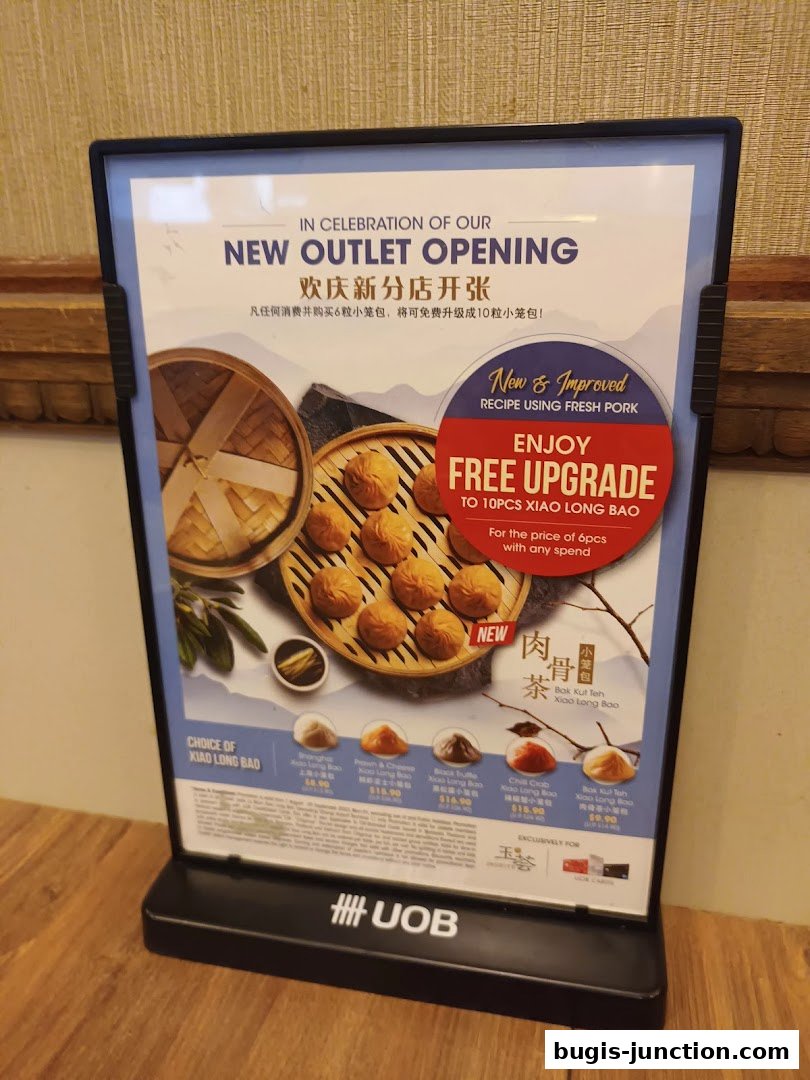 A promotional sign for Crystal Jade La Mian Xiao Long Bao announcing a new outlet opening and a free upgrade offer.