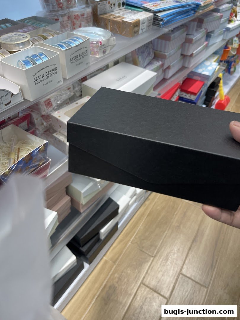A hand holds a black gift box in front of shelves stocked with various craft and gift items.