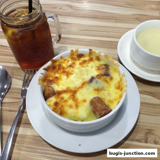 A close-up of a cheesy baked dish with sausages, served with iced tea and soup.