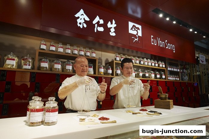 Two men in white coats weigh traditional Chinese medicine ingredients at a counter.