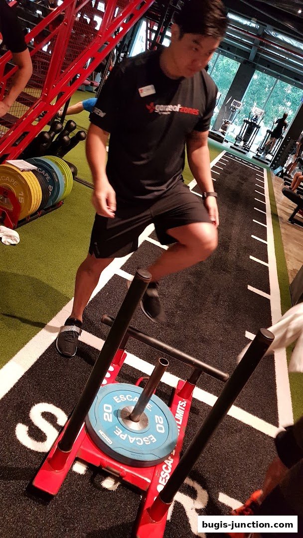 A fitness instructor demonstrates using a sled push machine with weight plates.