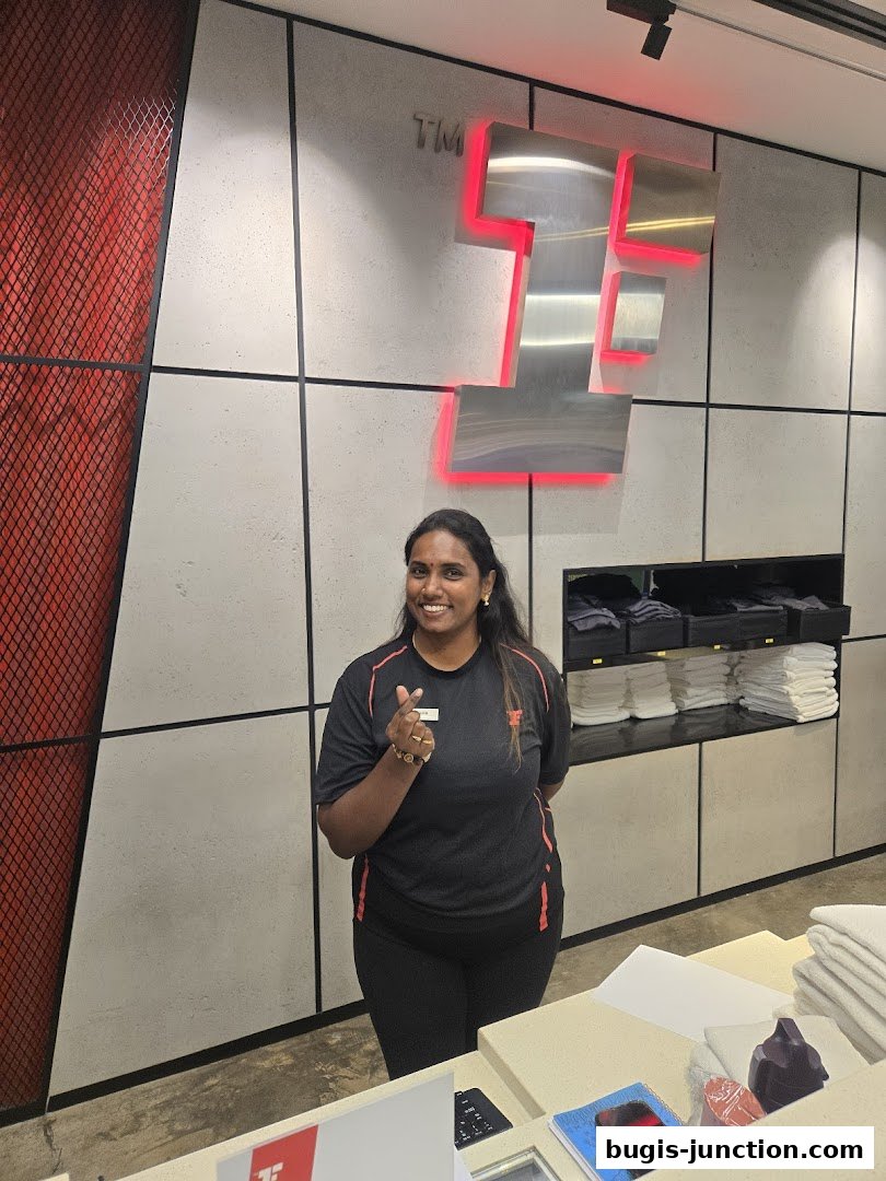 A smiling woman in a black t-shirt and leggings stands in front of a wall with a large illuminated logo.