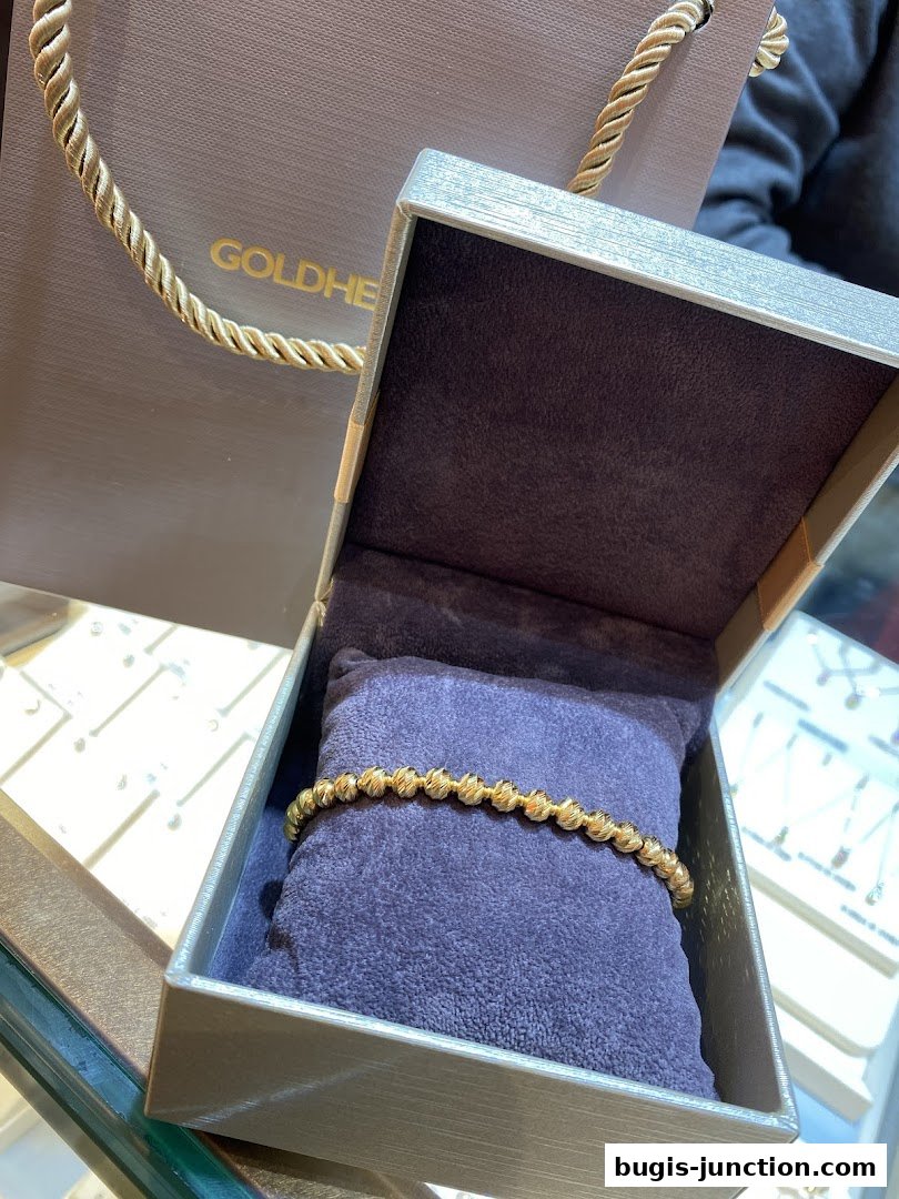 A gold beaded bracelet displayed in an open jewelry box with a shopping bag in the background.