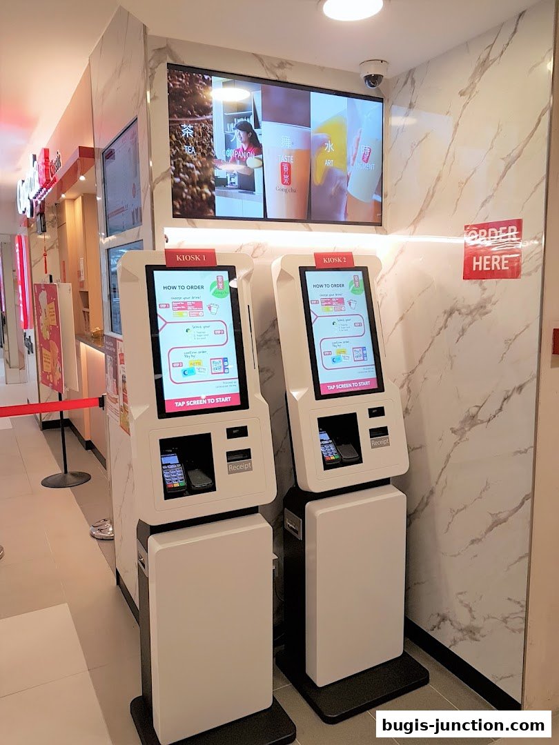 Two self-order kiosks are set up in front of a digital display at Gong Cha Classic.