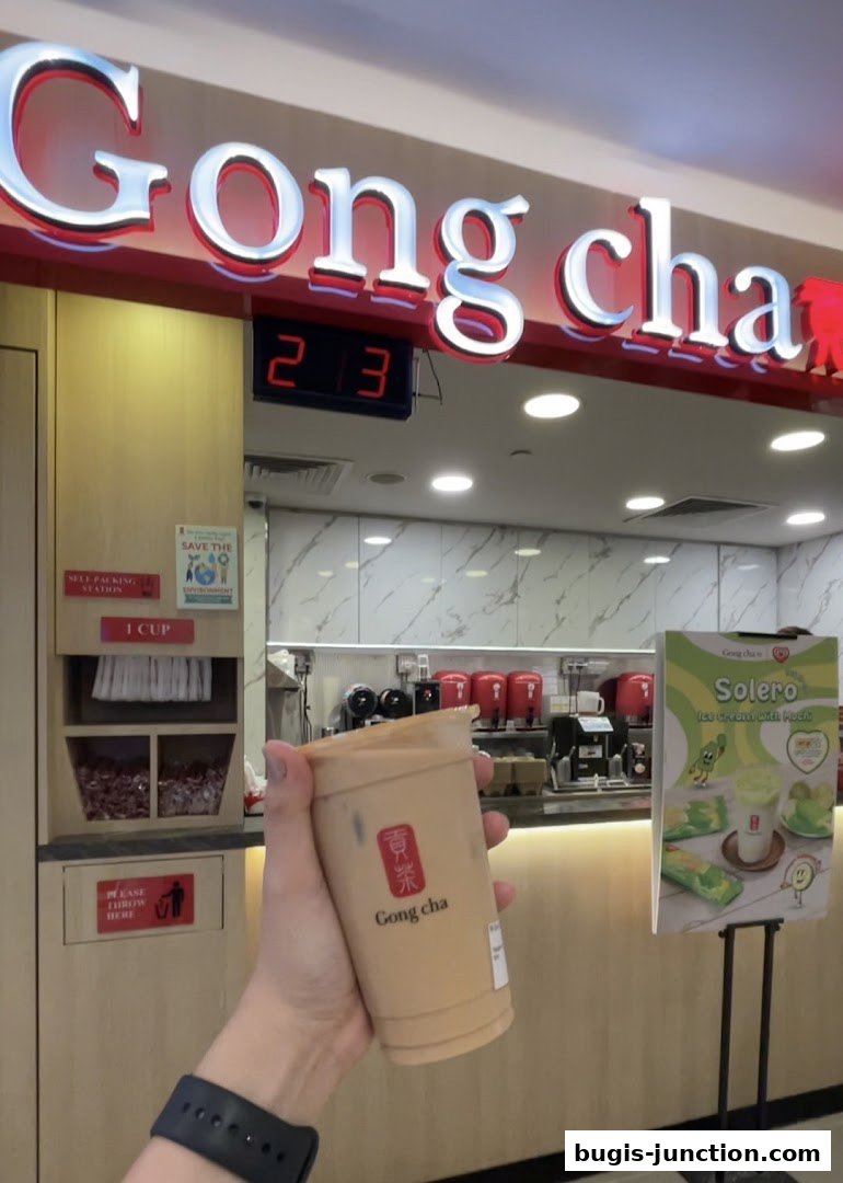 A hand holds a Gong Cha milk tea in front of the shop's illuminated sign.
