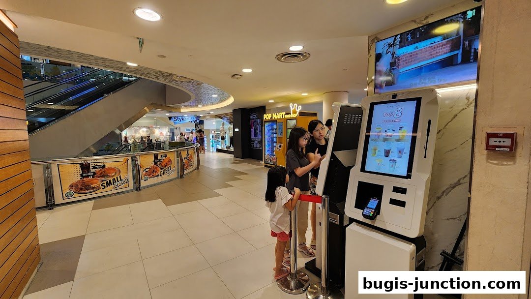 People ordering at a self-service kiosk in a mall with escalators and other shops in the background.