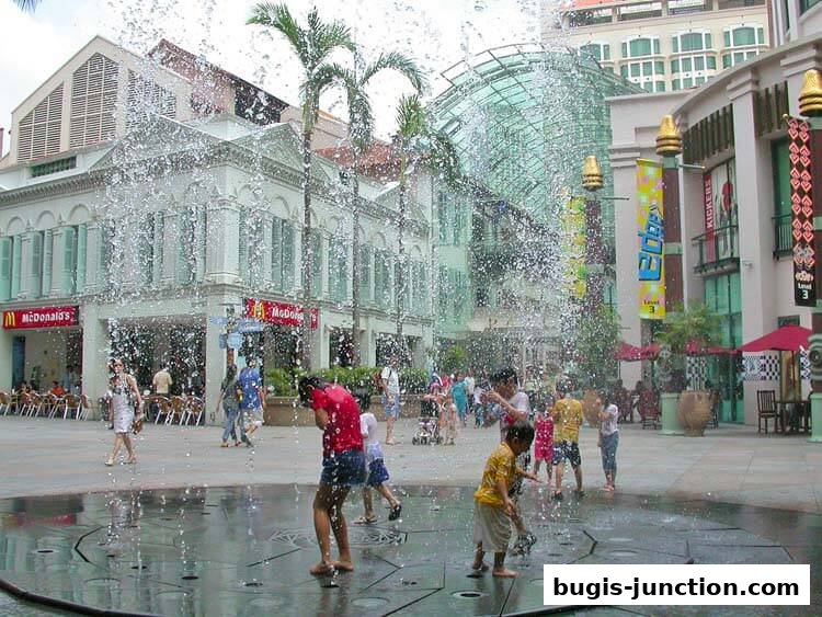 Children play in a fountain in front of shops and restaurants on a sunny day.