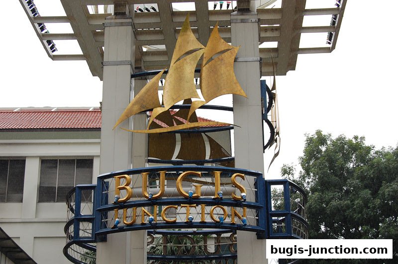 A sign for Bugis Junction with a ship sculpture and surrounding architecture.