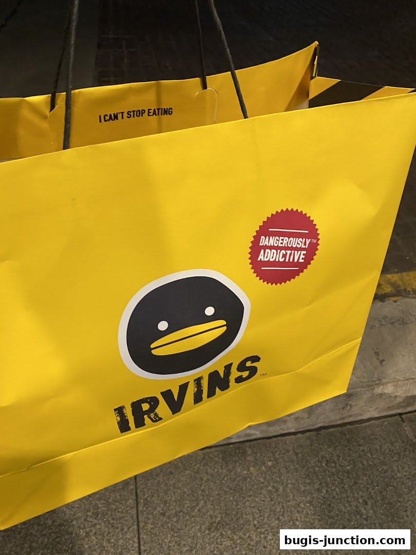 A bright yellow IRVINS shopping bag with a cartoon duck logo and 'Dangerously Addictive' slogan.