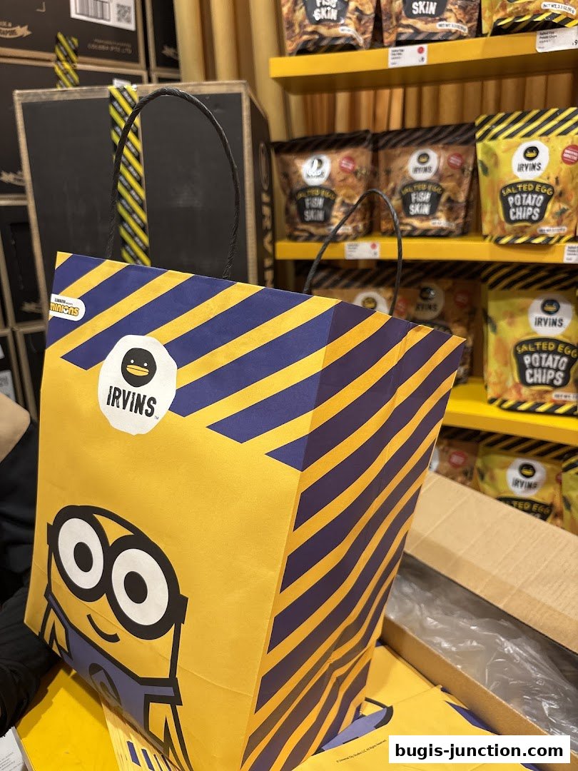 A shopping bag with a Minion character and IRVINS branding, with shelves of snacks in the background.
