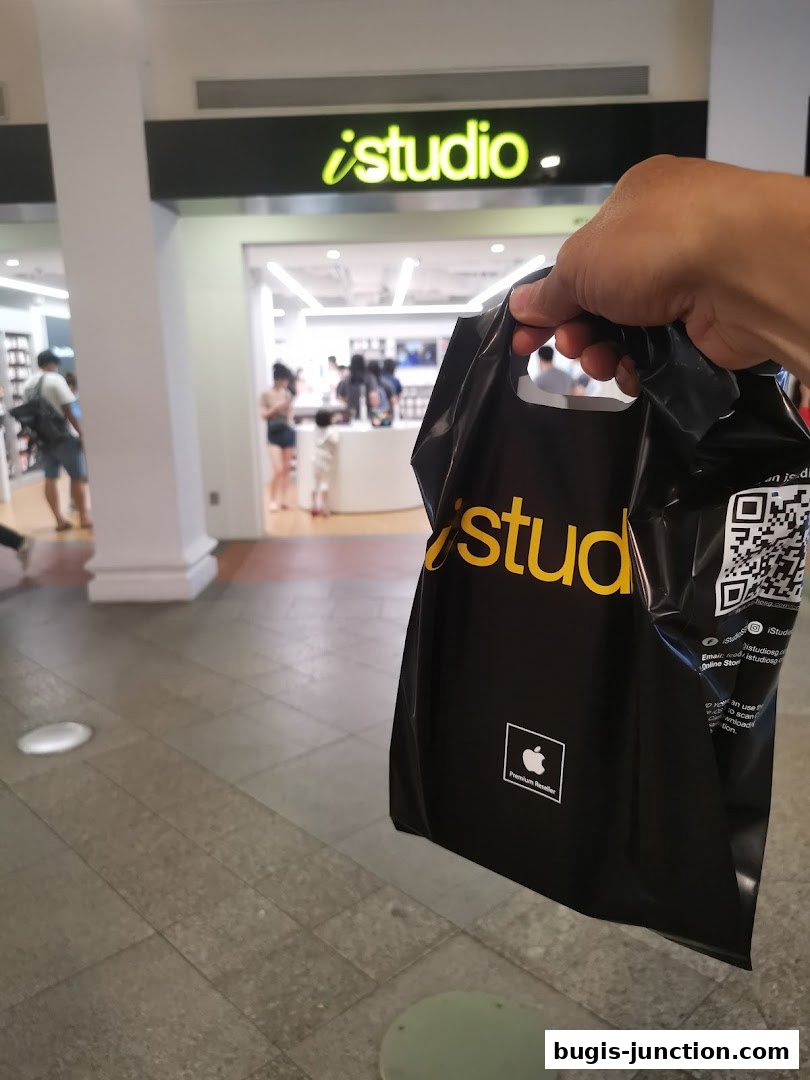 A hand holds a black shopping bag in front of the iStudio Bugis Junction store.