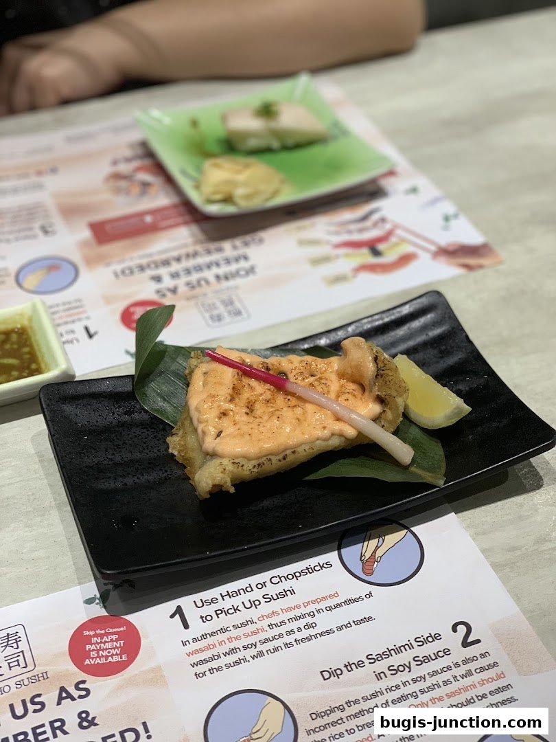A close-up of a sushi dish with a creamy sauce and a lemon wedge, served on a black plate.