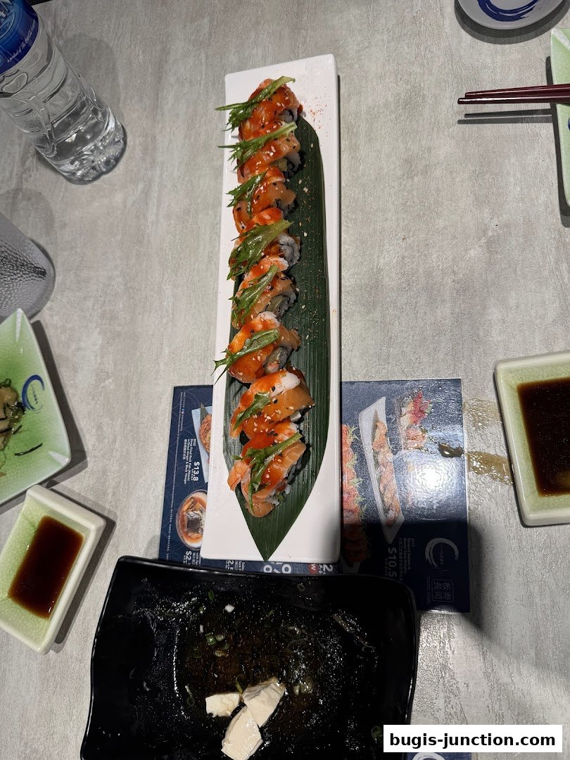 A long sushi roll with sauce and greens is served on a white plate with a banana leaf.