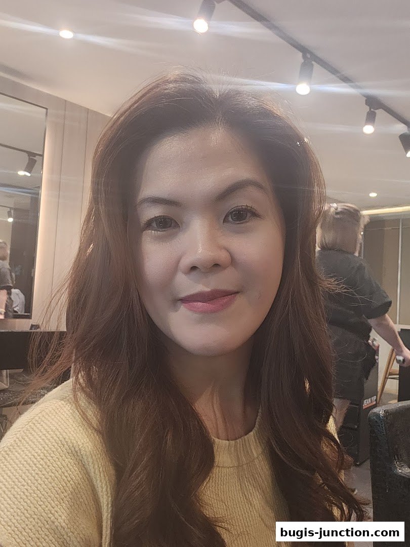 A woman with long, wavy brown hair smiles at the camera in a salon.