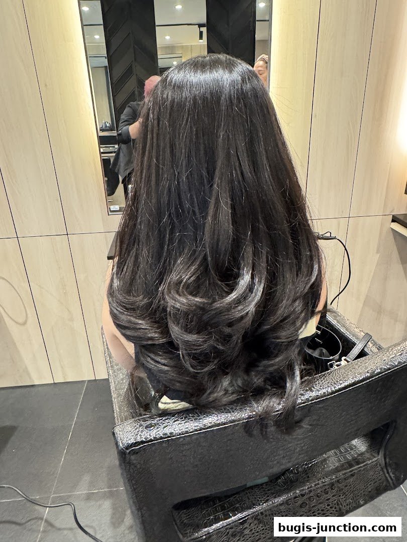 A person's long, dark, styled hair is shown from behind in a salon chair.