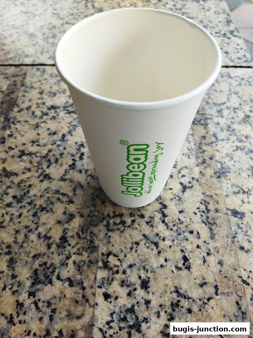 A white paper cup with the Jollibee logo and slogan on a speckled countertop.