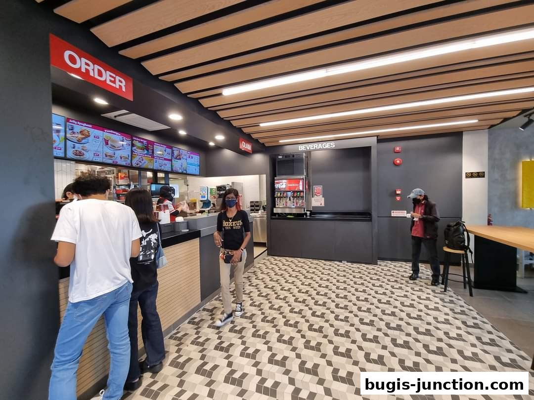 Customers queue at the order counter of a Jollibee restaurant with digital menus and a beverage station.