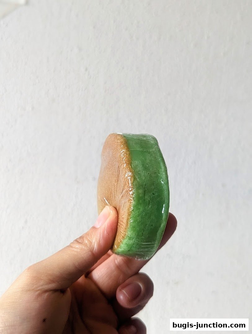 A hand holds a wrapped, round pastry with brown and green layers.