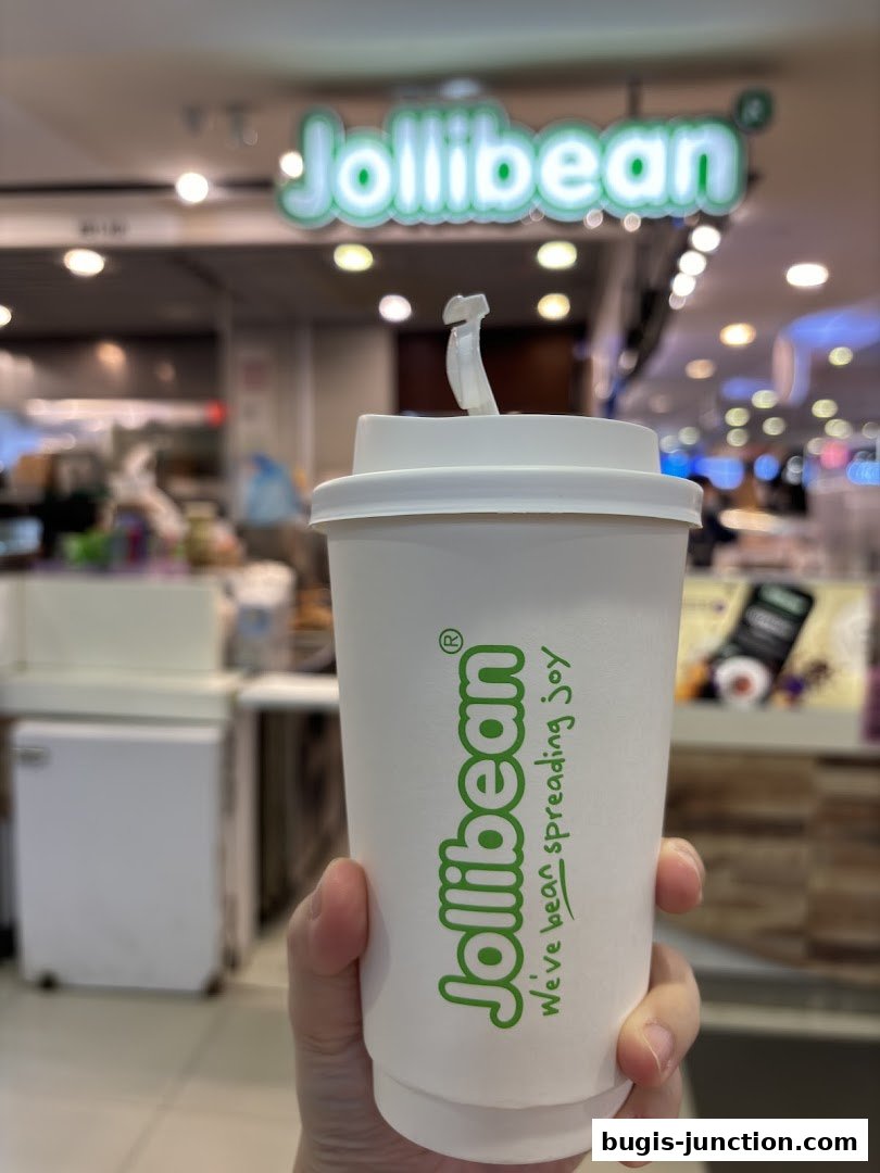 A hand holds a Jollibee branded cup with the store's logo and slogan in the background.