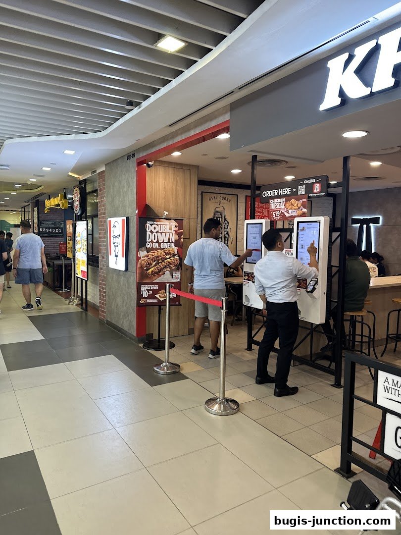 KFC restaurant with customers ordering at self-service kiosks and a promotional poster.