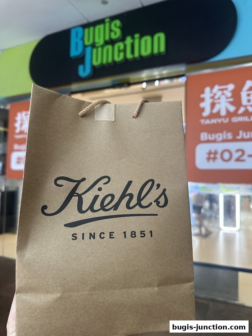 A Kiehl's shopping bag is held up in front of the Bugis Junction mall signage.