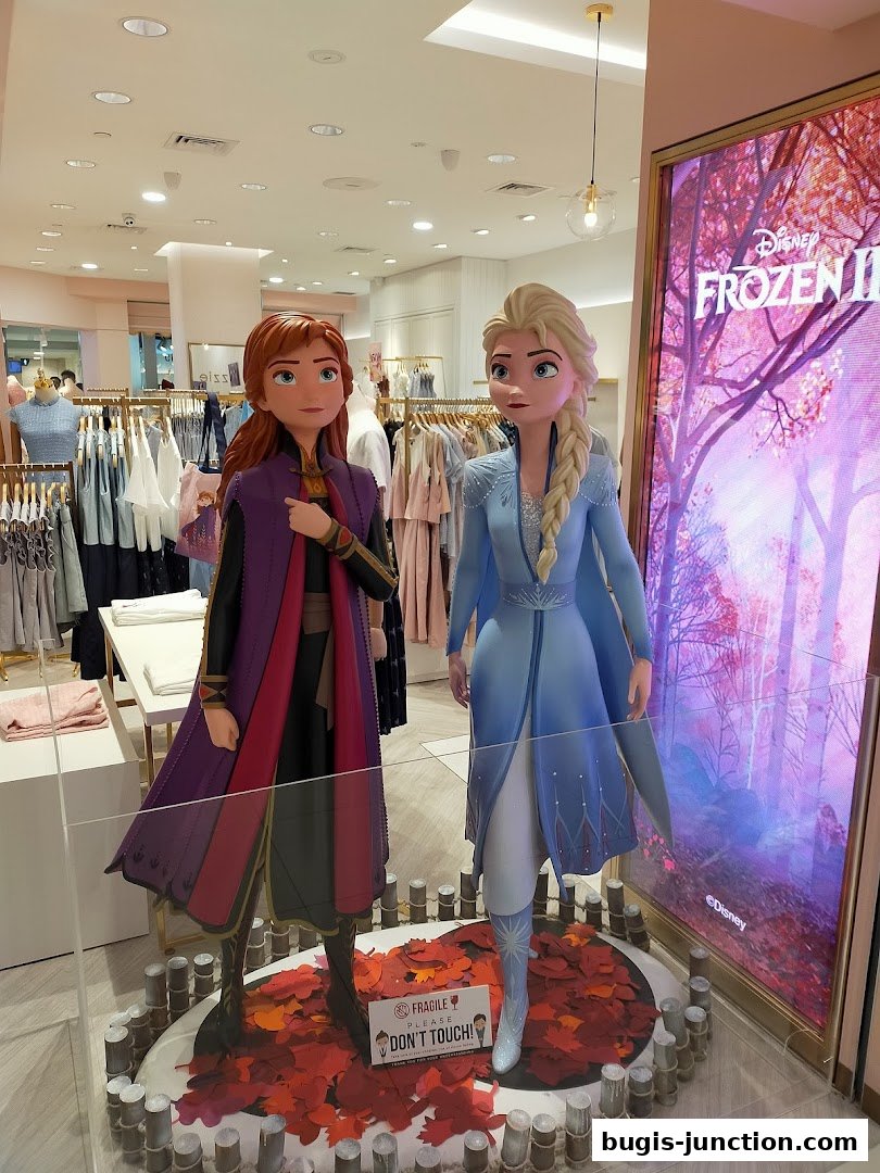 Anna and Elsa figures from Frozen II displayed in a shop with clothing racks and a digital screen.