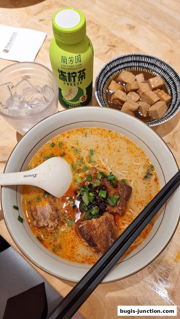 A bowl of hearty noodle soup with tender meat, a side dish of braised tofu, and a bottle of iced tea.