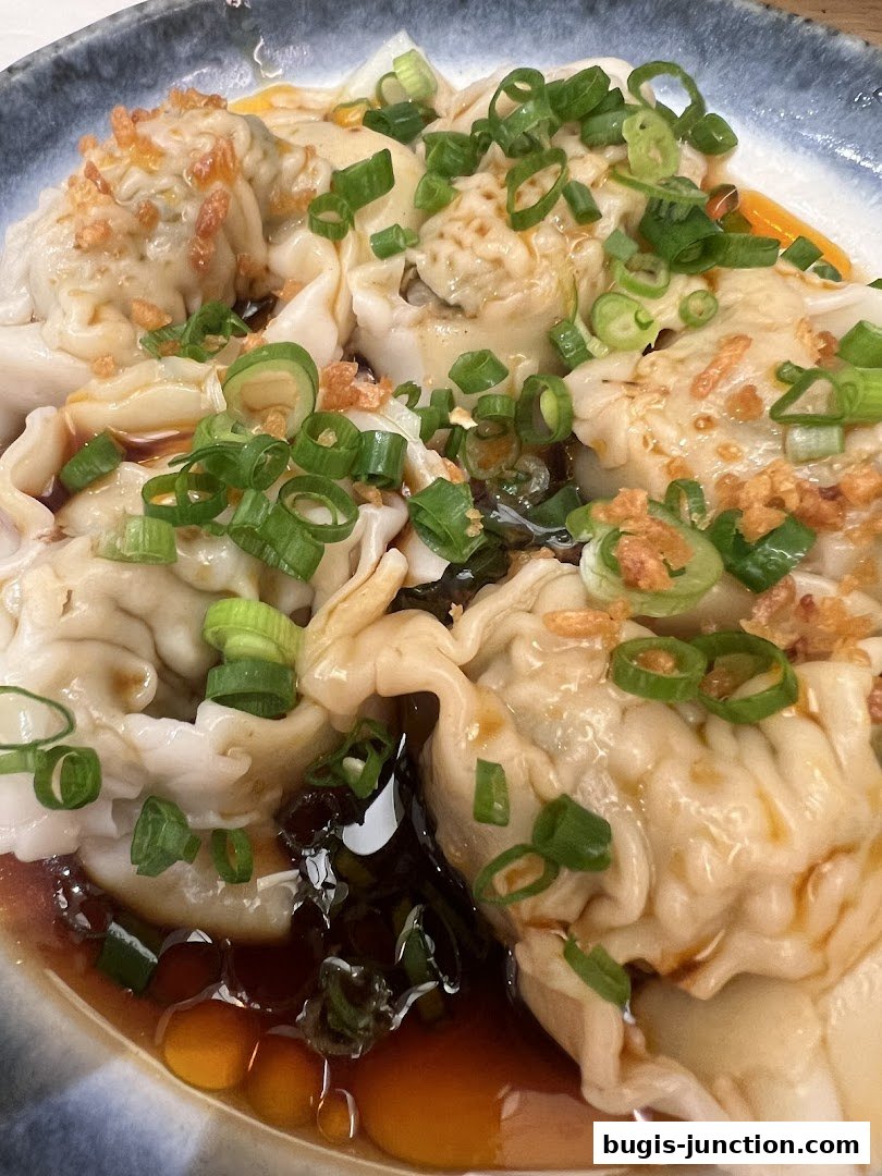 A close-up shot of delicious wontons in a savory broth, garnished with chopped scallions and crispy garlic.